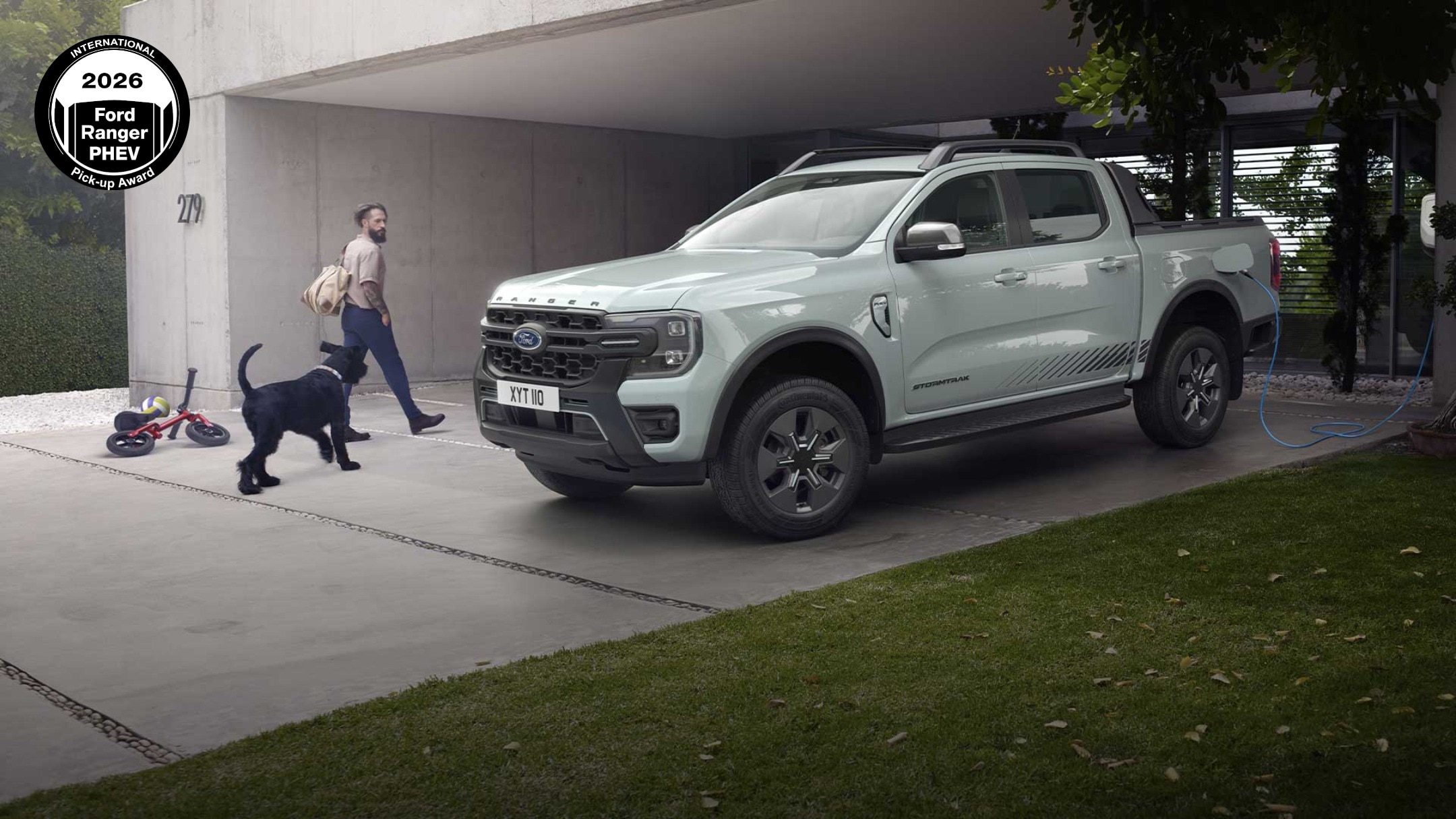Un uomo e un cane si dirigono verso un pick-up Ford Ranger ibrido plug-in grigio mentre si ricarica in un vialetto. 