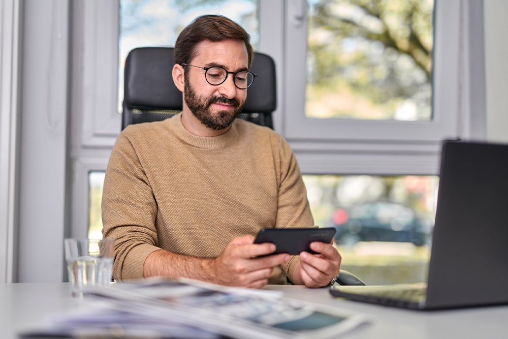 Persona seduta a una scrivania, guarda uno smartphone che tiene in mano, con un laptop sulla scrivania.