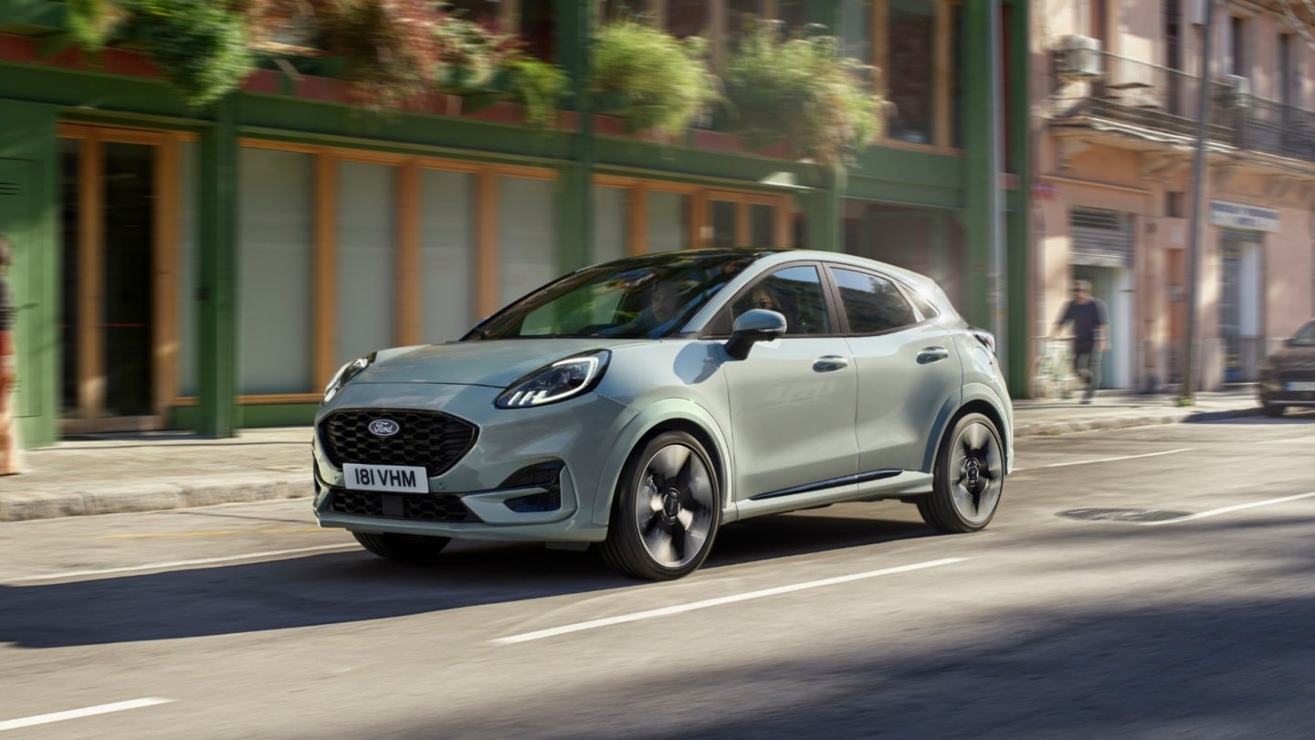 Una Ford Puma grigio chiaro sfreccia su una strada urbana, con lo sfondo sfocato che accentua il movimento.