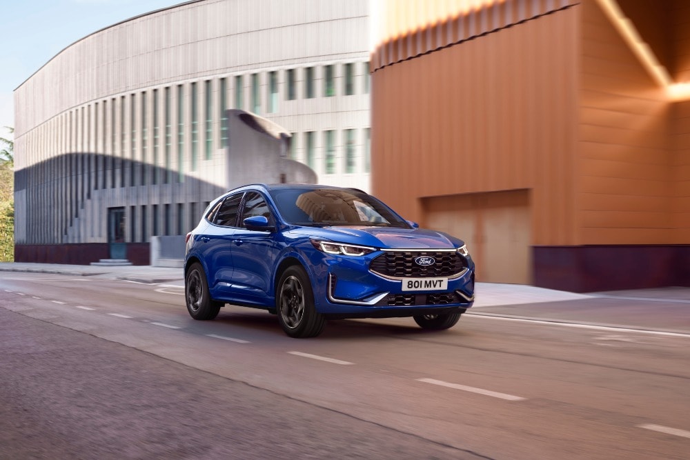 Ford Kuga blu che guida su una strada cittadina con edifici moderni sullo sfondo.