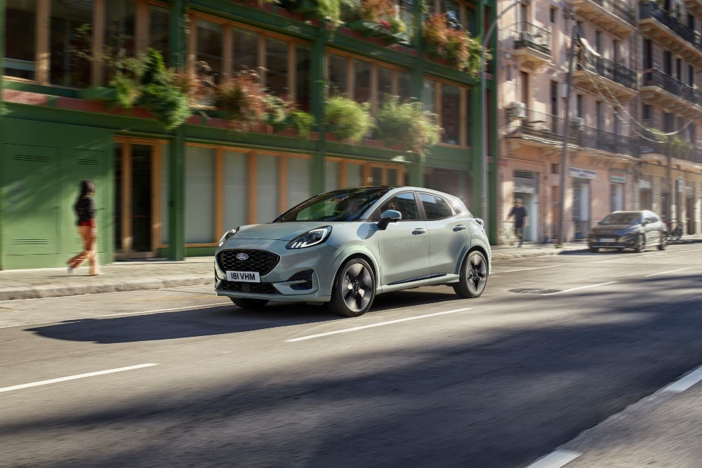 Ford Puma verde chiaro che guida su una strada cittadina con edifici e una persona che cammina sullo sfondo.