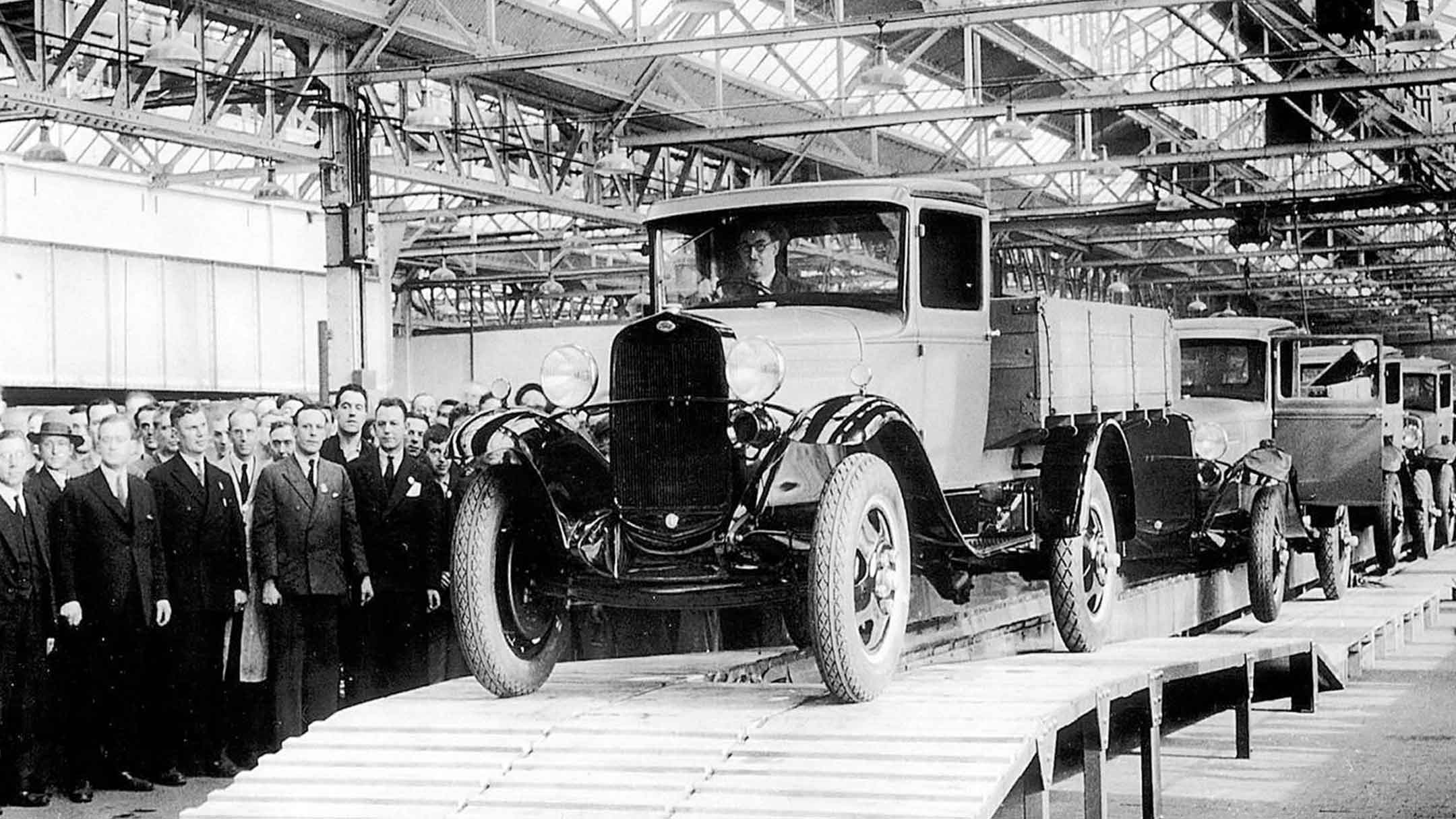 Historical black and white picture of a car factory with the first Ford vehicle on an assembly line.