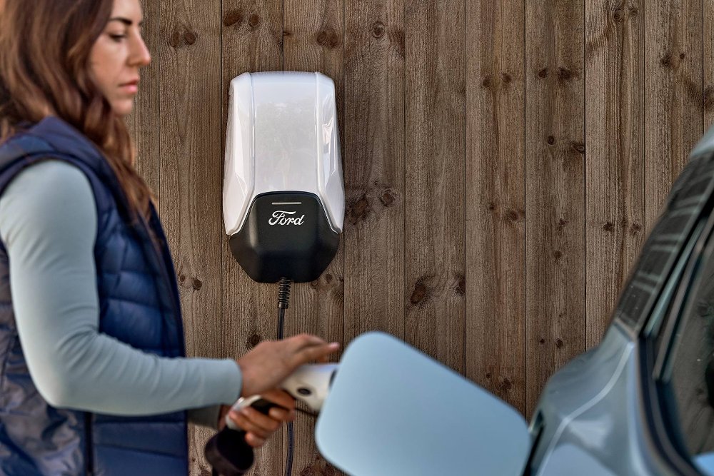 Una donna collega la sua Ford Explorer elettrica blu a una stazione di ricarica domestica per veicoli elettrici.