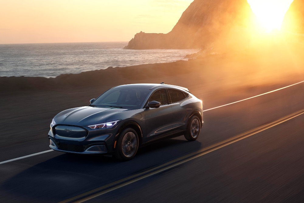 Un'auto elettrica Ford Mustang Mach-E percorre una strada costiera al tramonto.