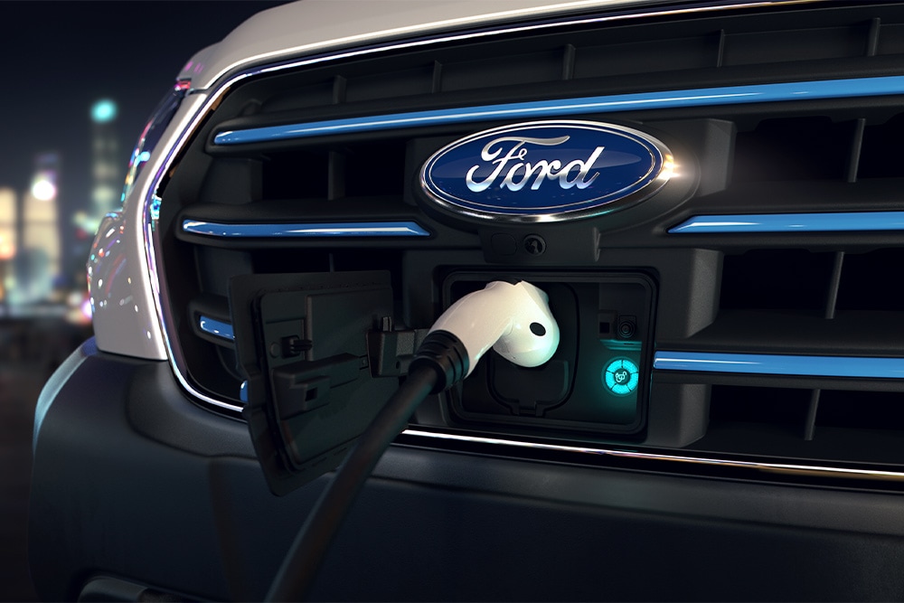Primo piano di un punto di ricarica del furgone Ford E-Transit e dello stemma ovale blu Ford sulla griglia anteriore.
