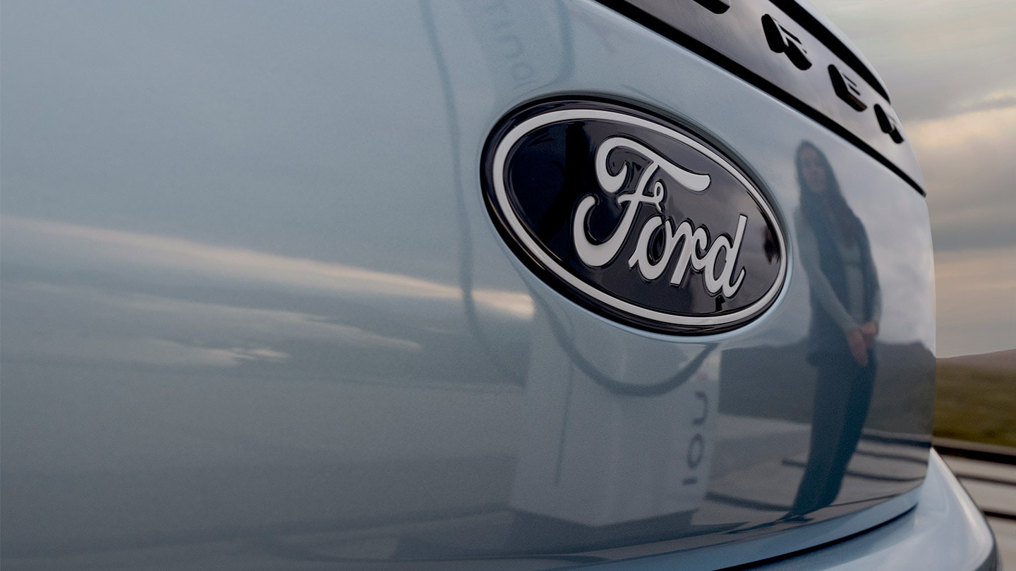 Logo Ford su una Ford Explorer elettrica blu con una stazione di ricarica pubblica riflessa nell'auto.