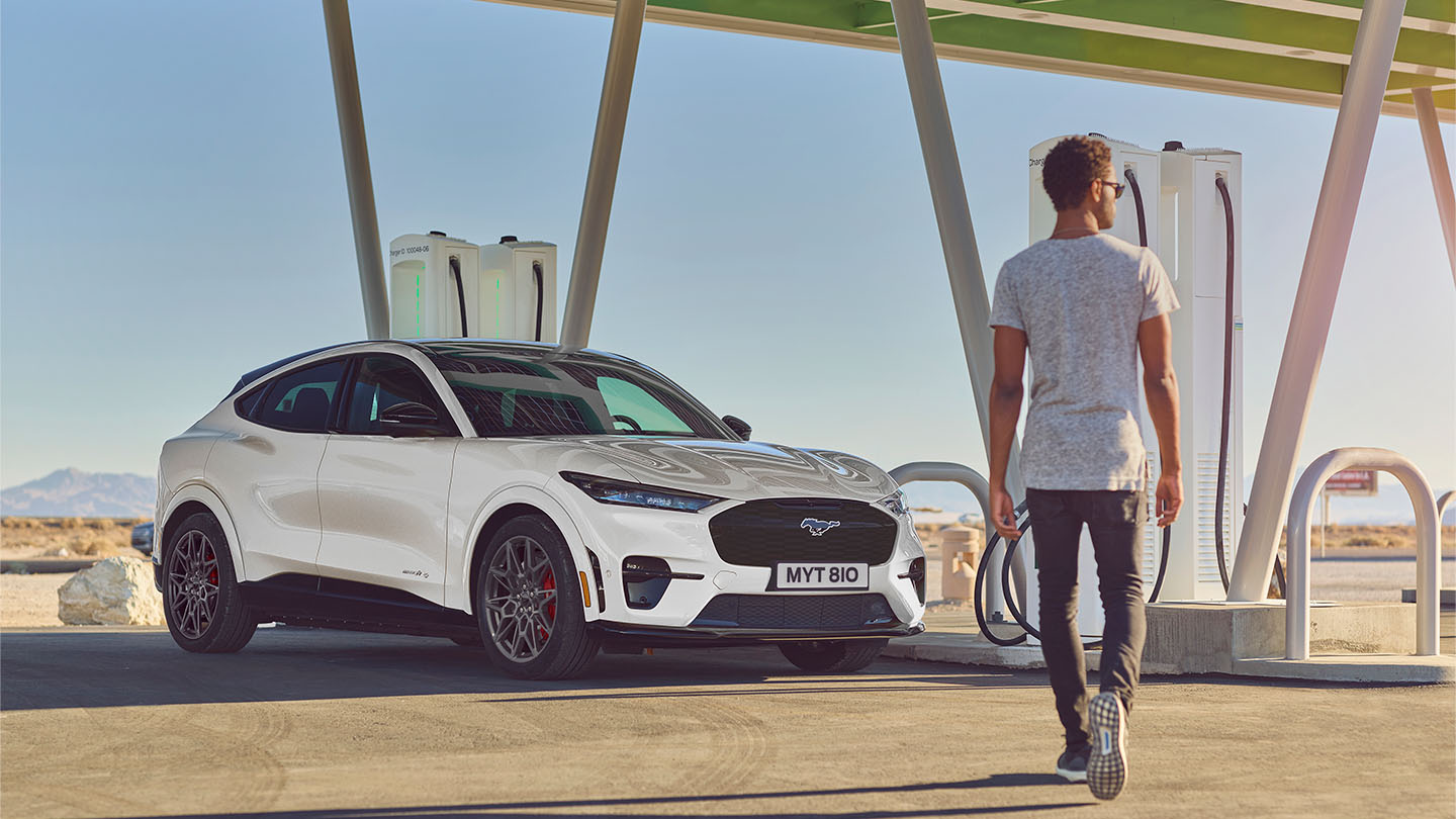 Un Ford Mustang Mach-E bianco in una stazione di ricarica pubblica. Un uomo con maglietta bianca cammina verso il veicolo.