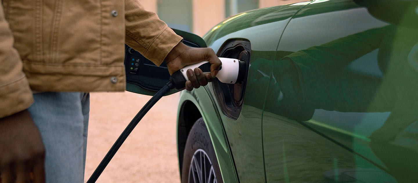 Una persona con giacca beige che collega un cavo di ricarica elettrico al suo Ford Mustang Mach-E elettrico verde.