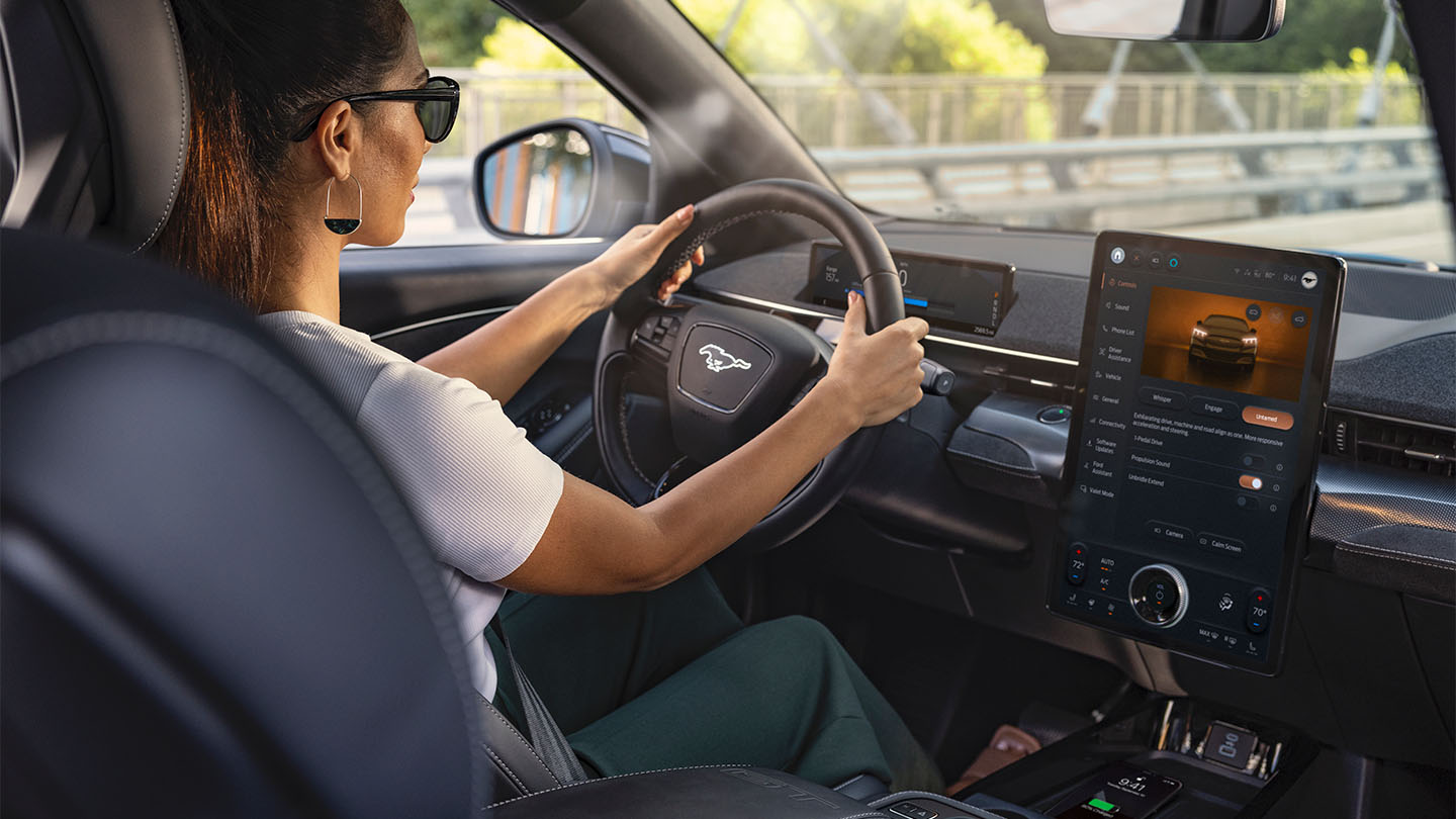 Una donna guida un Mustang Mach-E con le mani sul volante. Il touchscreen da 15,5" mostra i dati del veicolo.