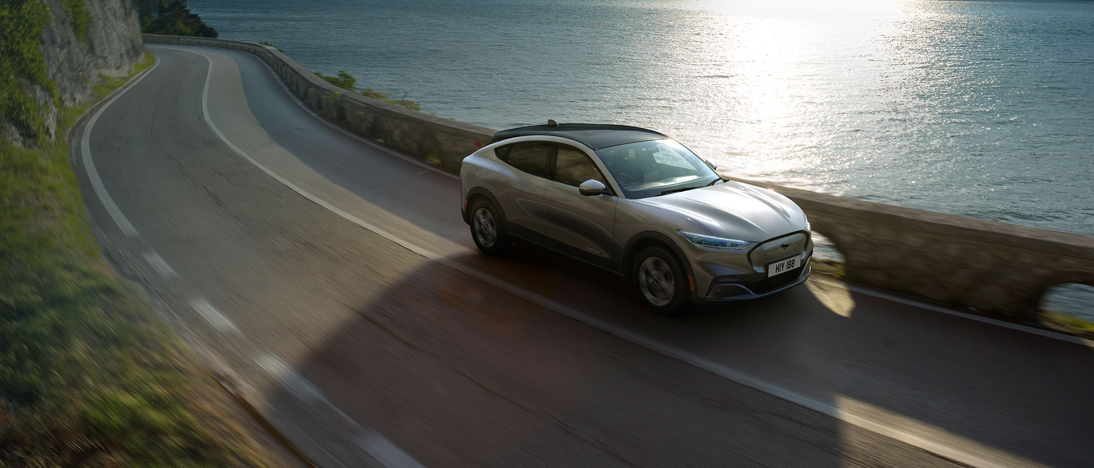 Ford Mustang Mach-E All Electric grigia in movimento che costeggia scogliere soleggiate e mare