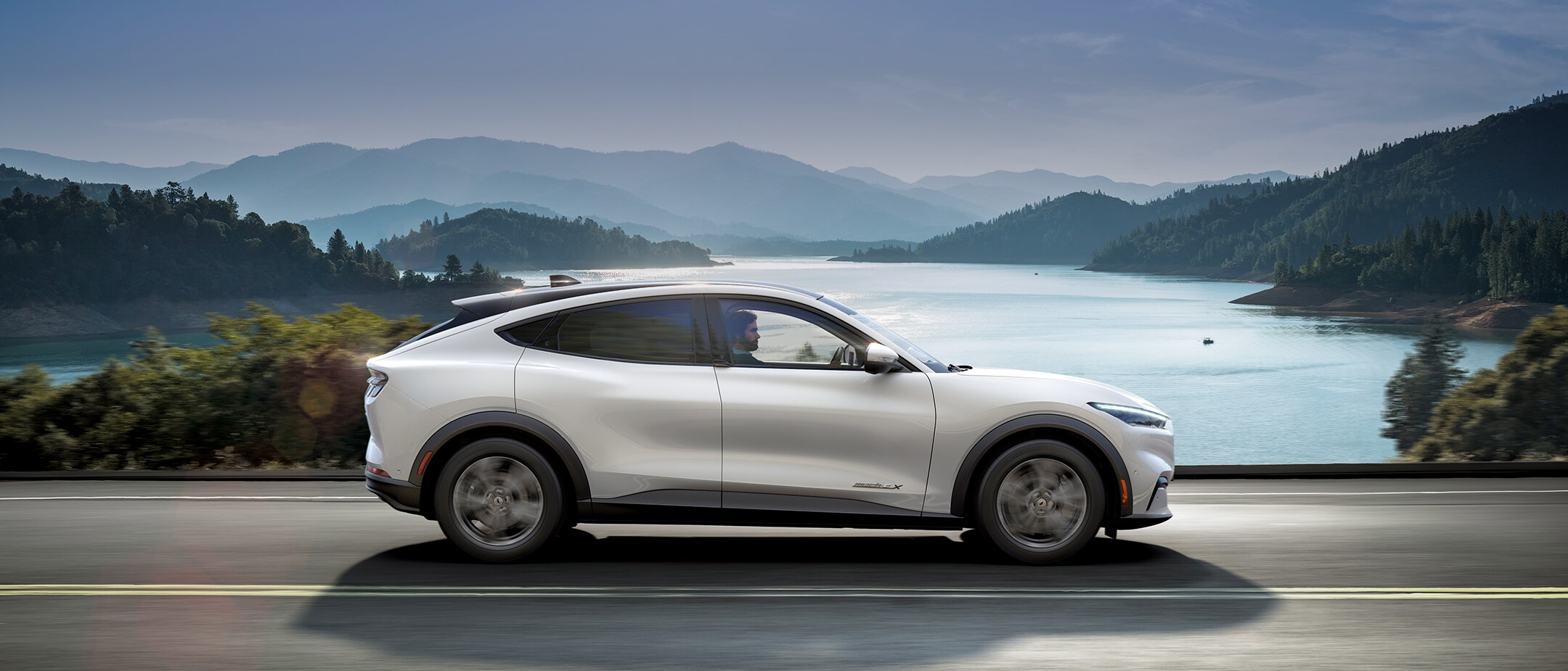 Ford Mustang Mach-E All Electric grigia in movimento che costeggia scogliere soleggiate e mare