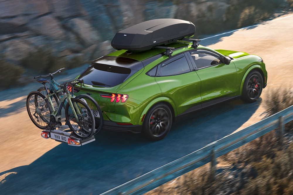 Vista posteriore di una Ford Mustang Mach-E verde con box da tetto e portabiciclette mentre percorre una strada panoramica.