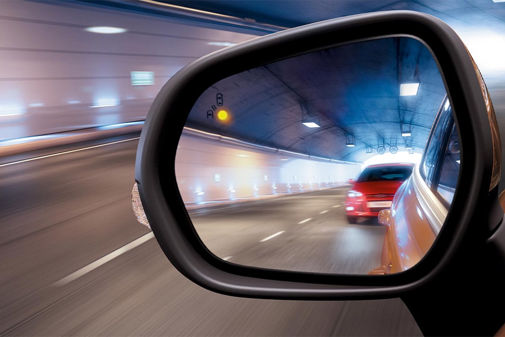 Un primo piano dello specchietto retrovisore laterale in un tunnnel, che mostra un'auto rossa che si avvicina nell'angolo cieco del conducente.