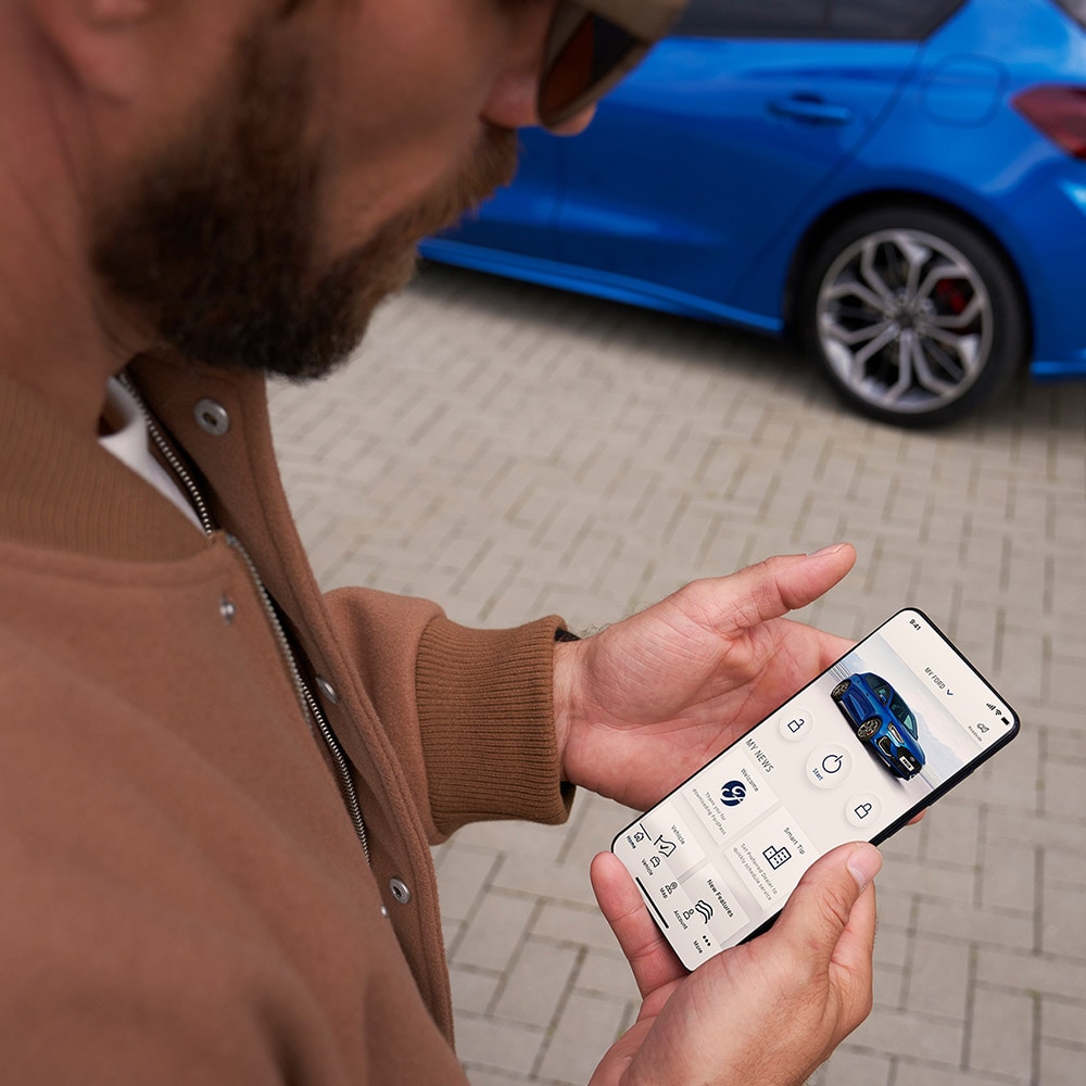 Una persona con in mano uno smartphone che visualizza l'app FordPass™, con una Ford Focus blu sullo sfondo.
