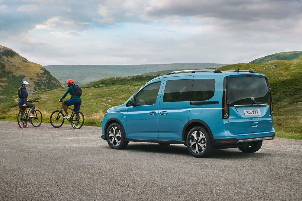 Un Ford Tourneo Connect blu è parcheggiato su una strada tra colline verdi, con due ciclisti vicini.