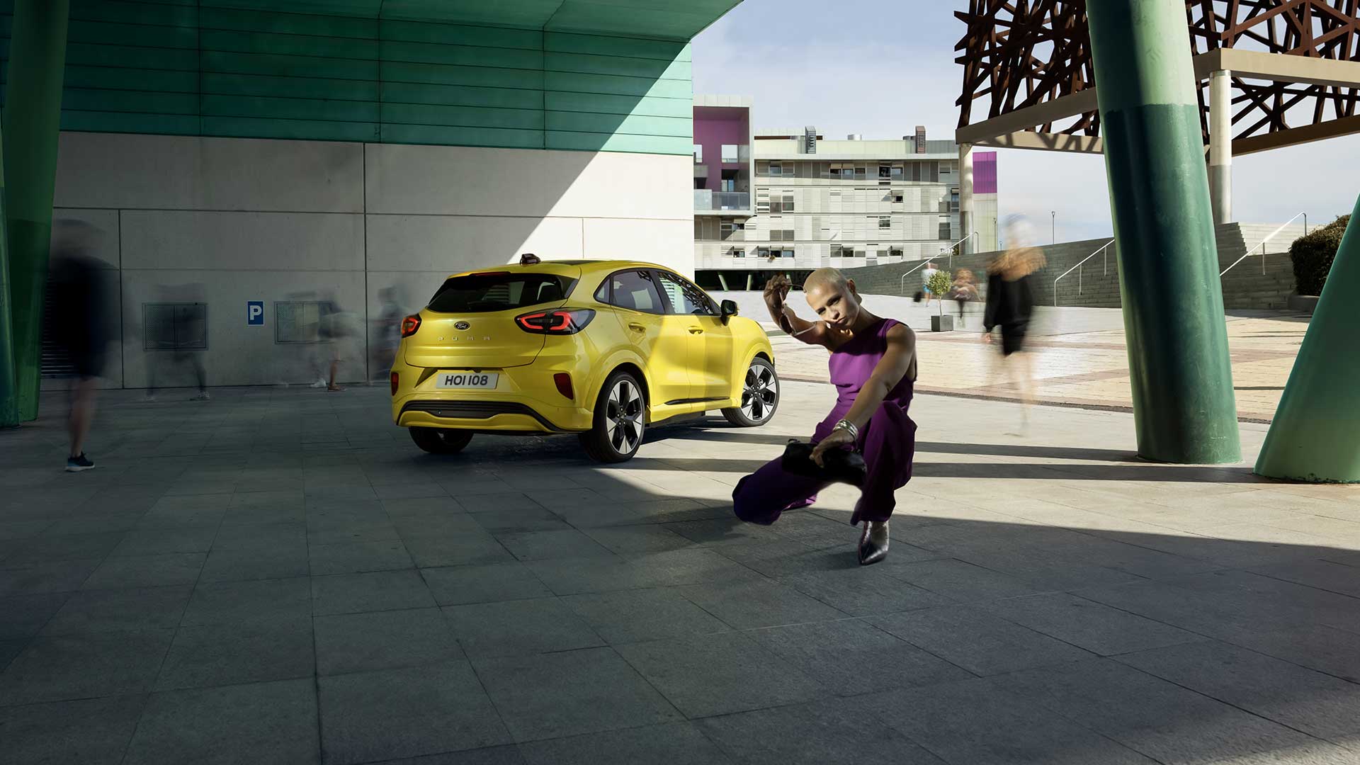 Vista posteriore di un Ford Puma Gen-E® elettrico giallo parcheggiato sotto un ponte, accanto a una persona vestita di viola.