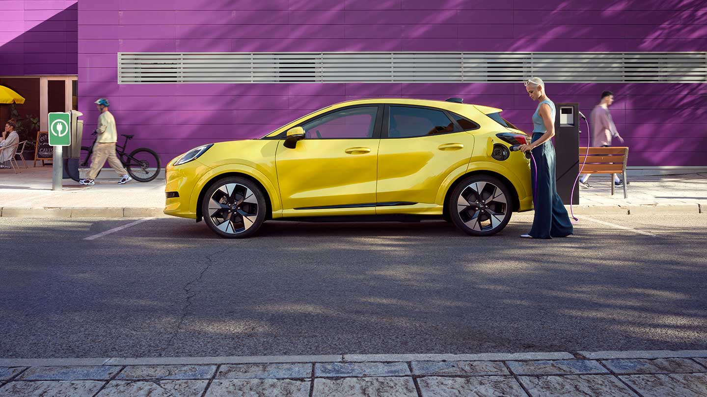 Vista laterale del Ford Puma Gen-E® giallo collegato a una stazione di ricarica pubblica all'interno di un parcheggio.