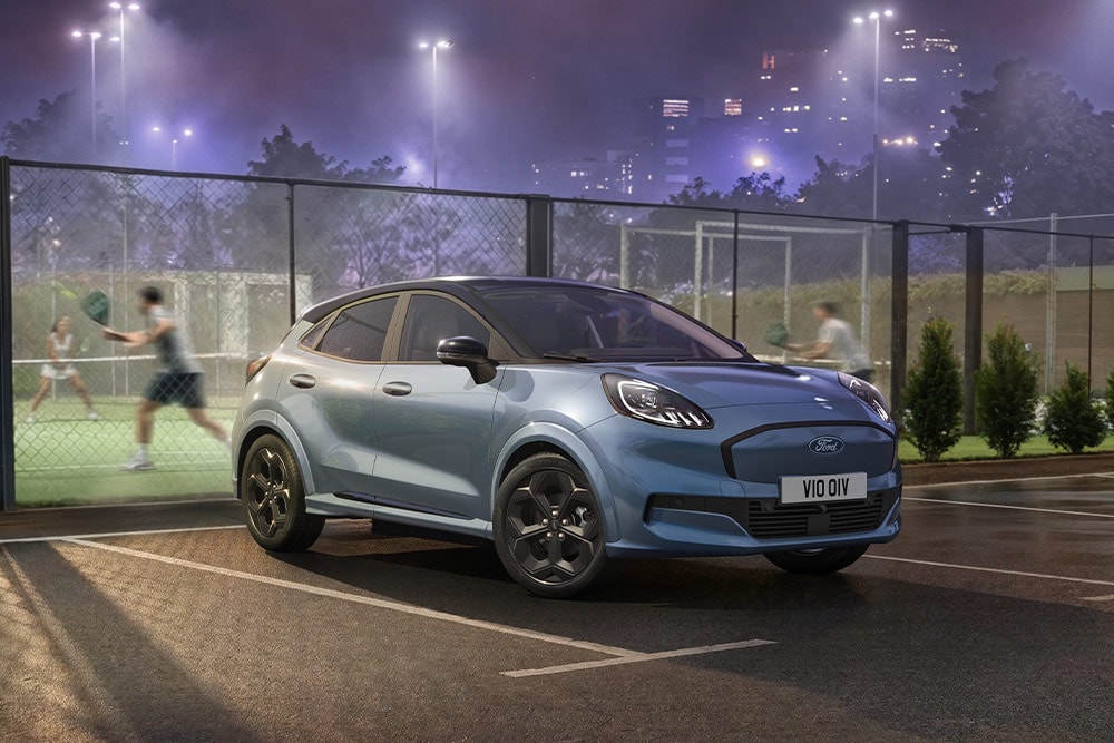 Una Ford Puma Gen-E blu blu parcheggiata accanto a un campo da tennis, con le luci della città sullo sfondo.