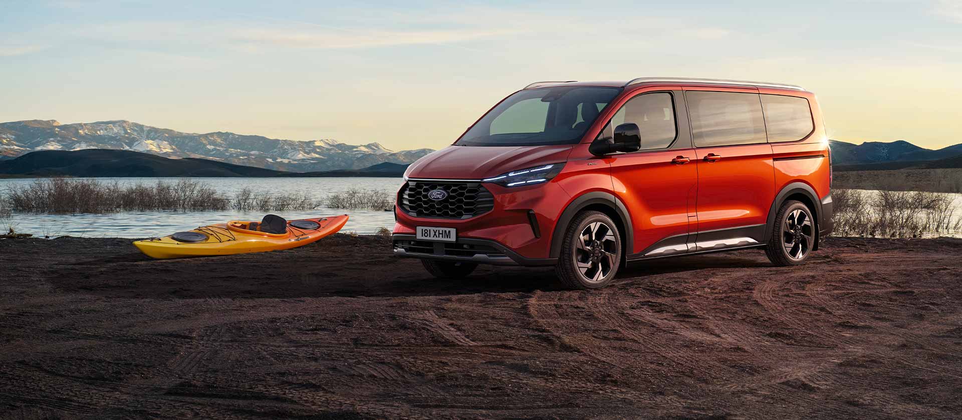Un furgone Ford Tourneo Custom rosso parcheggiato nelle vicinanze di un lago con un kayak. 