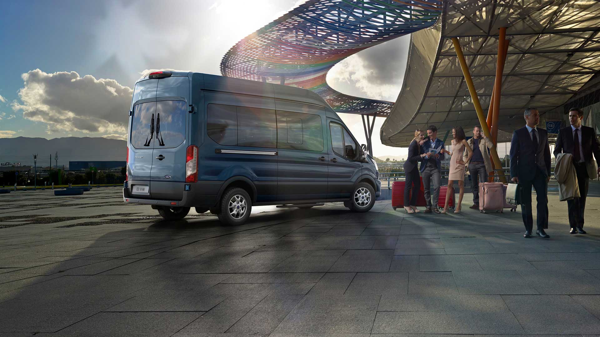 Rear 3/4 view of a blue Ford Transit Minibus parked at an airport, with people and luggage nearby.
