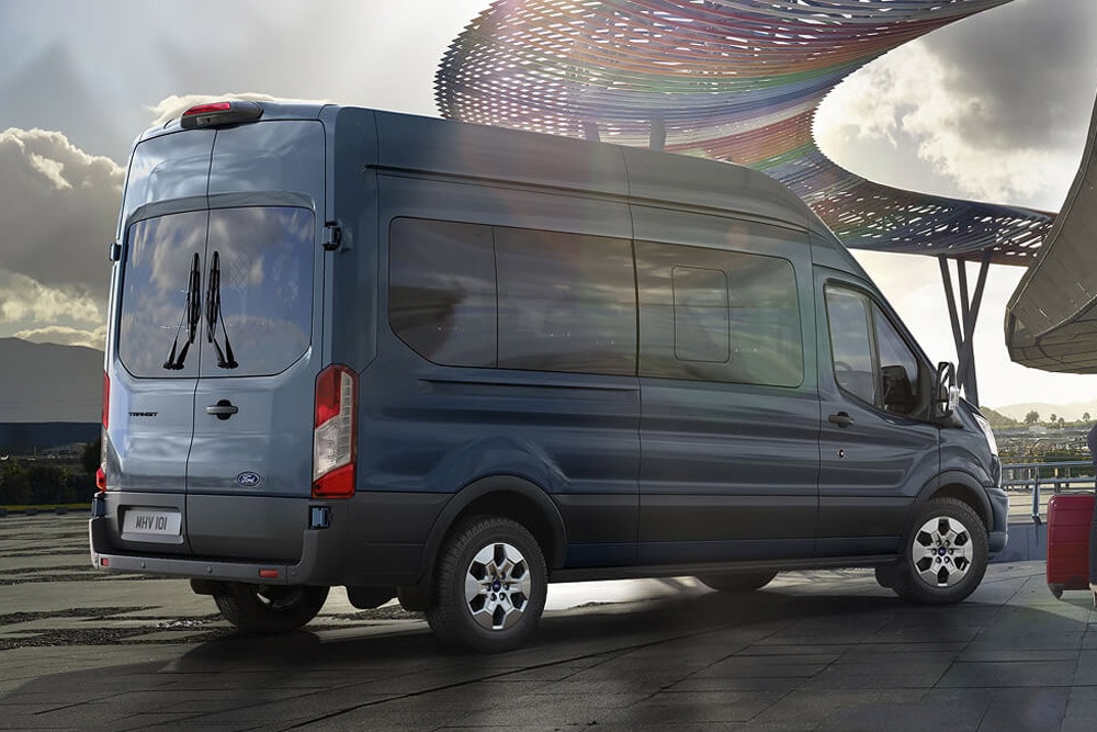 Rear 3/4 view of a blue Ford Transit Minibus parked outdoors, with a modern, colorful structure in the background.