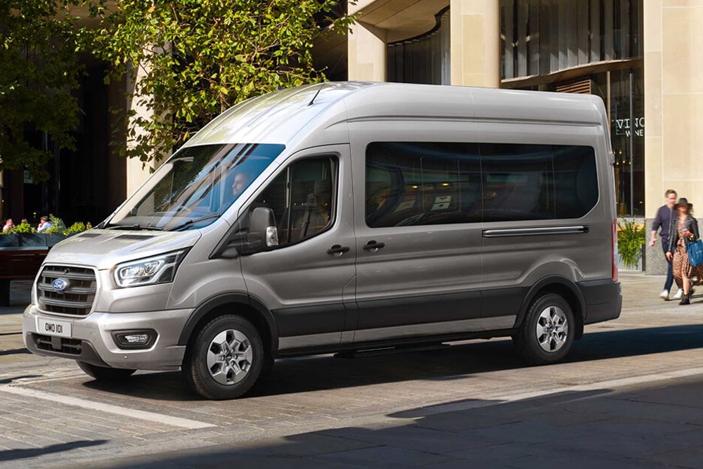 Side 3/4 view of a silver Ford Transit Minibus parked on a city street with people walking in the background.