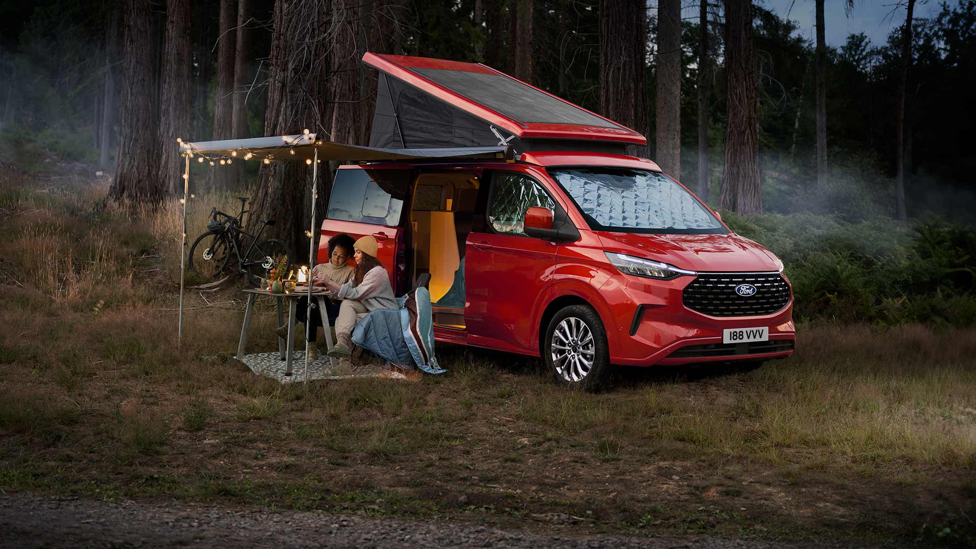 Due donne sono sedute sotto una tenda da sole, accanto alle porte laterali aperte di un Ford Transit Custom Nugget rosso, con il tetto ribaltabile alzato.