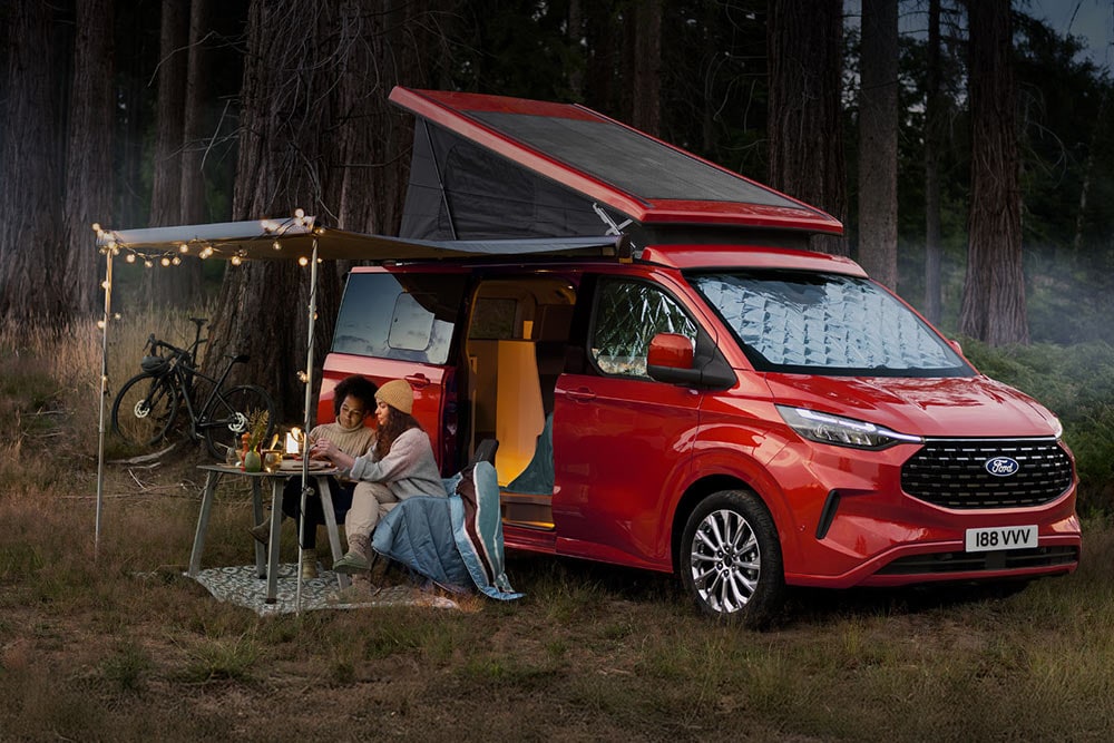 Due donne sono sedute sotto una tenda da sole, accanto alle porte laterali aperte di un Ford Transit Custom Nugget rosso, con il tetto ribaltabile alzato.