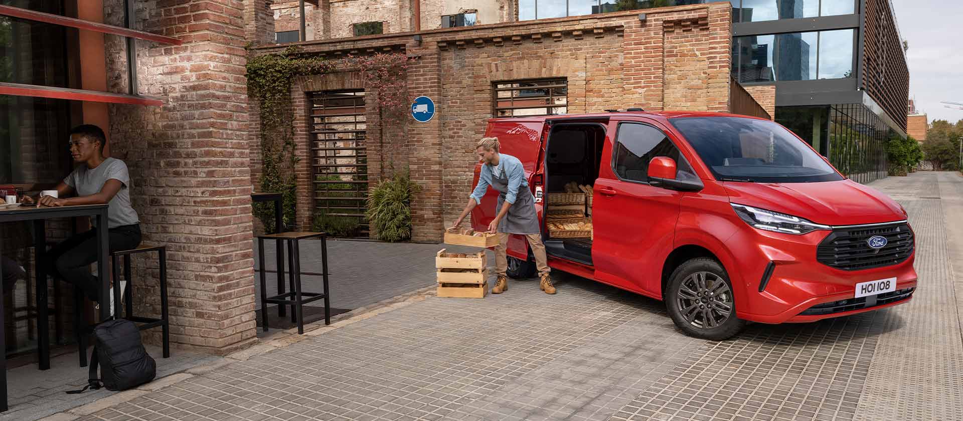 Una persona scarica delle casse da un furgone Ford Transit Custom rosso parcheggiato davanti a un bar. 