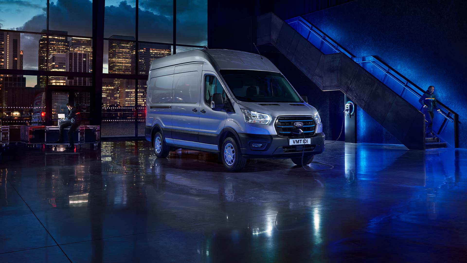 Un furgone elettrico E-Transit in versione argento parcheggiato al chiuso con una città visibile sullo sfondo attraverso grandi vetrate. 