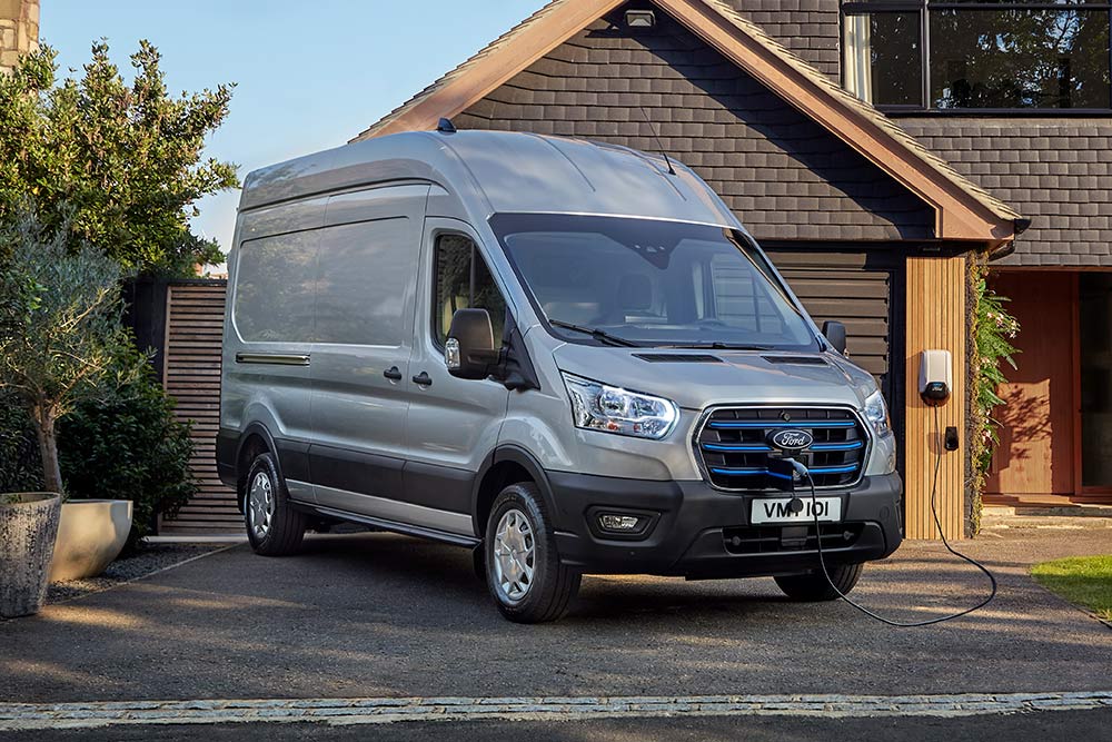 Un furgone Ford E-Transit  versione argento parcheggiato vicino a una casa, collegato a una stazione di ricarica.
