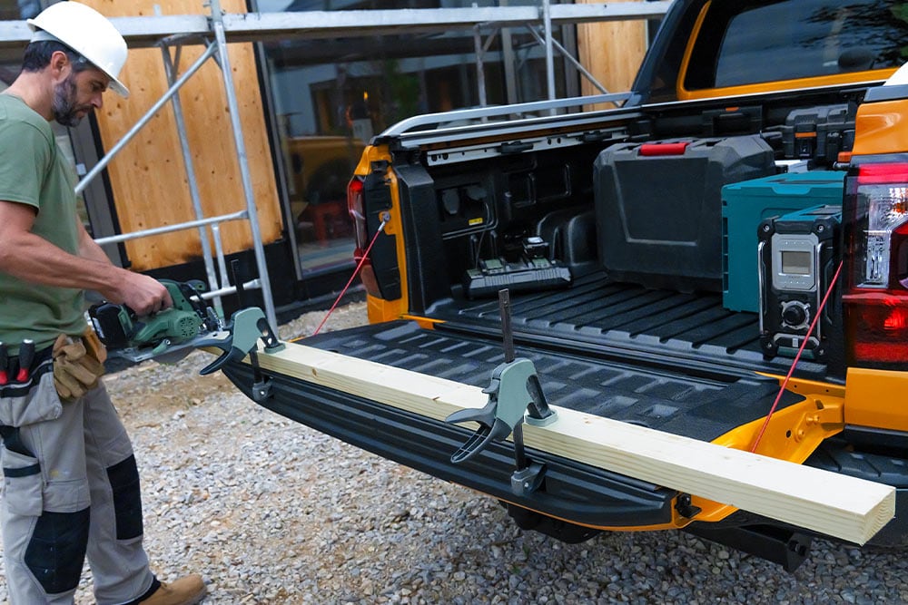 The tailgate of a yellow Ford Ranger is down, with a wooden plank secured to it using clamp.