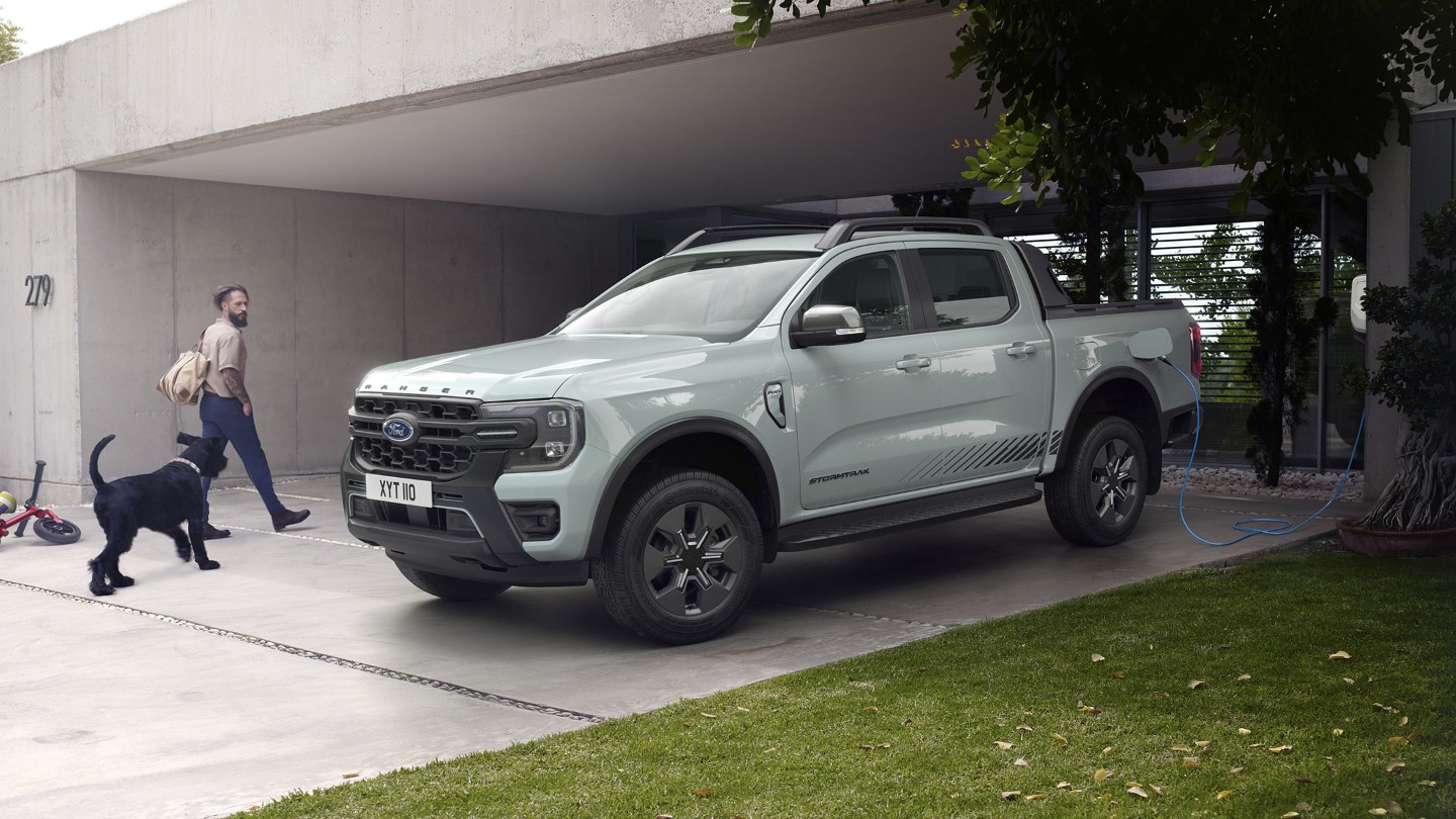 Un uomo e il suo cane entrano in una casa moderna, passando davanti a un Ford Ranger Stormtrak Plug-in Hybrid parcheggiato e in carica. 