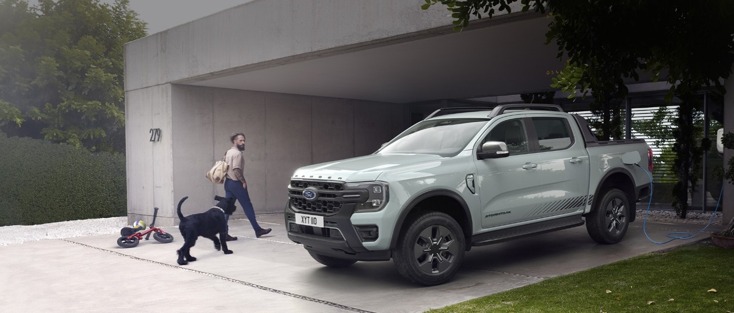 Un uomo e il suo cane entrano in una casa moderna, passando davanti a un Ford Ranger Stormtrak Plug-in Hybrid parcheggiato e in carica. 