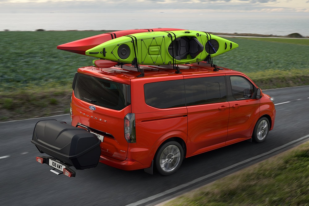 Ford Pro Transit rosso con due kayak sul tetto, che traina un portapacchi e guida lungo una strada costiera.