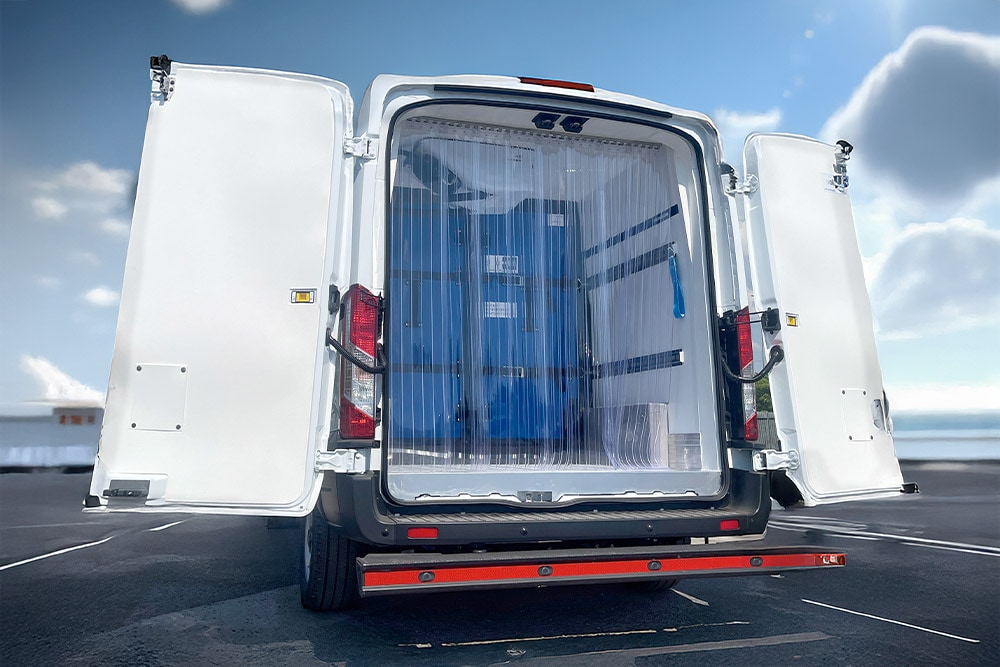 Vista posteriore di un furgone refrigerato Ford Transit bianco con porte aperte e tende a strisce di plastica.