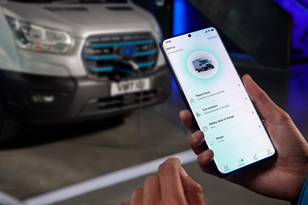 Una persona che tiene uno smartphone che mostra un'interfaccia di app, con un furgone Ford Pro Transit argentato sfocato sullo sfondo.