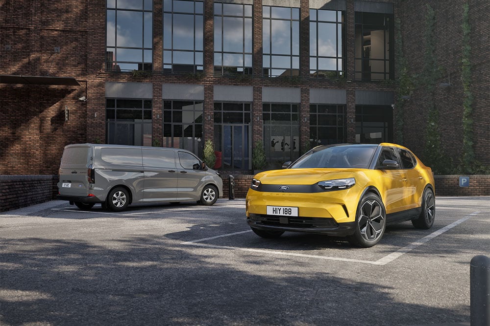 Un furgone Ford grigio e un Capri giallo parcheggiati di fronte a un moderno edificio in mattoni.