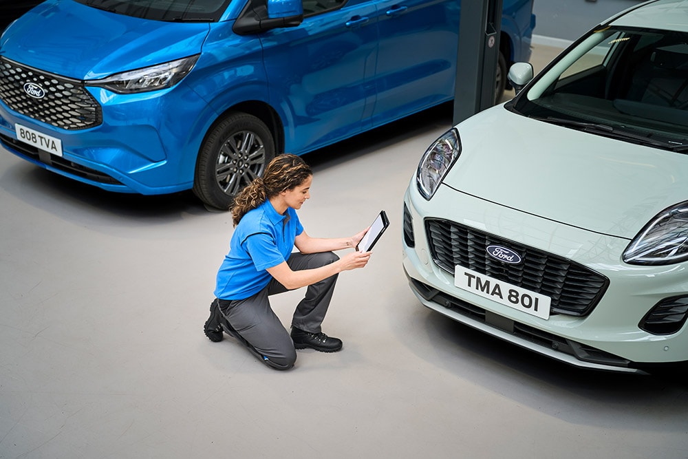 Un tecnico che guarda un tablet, inginocchiato tra un furgone Ford blu e una Ford Puma bianca.