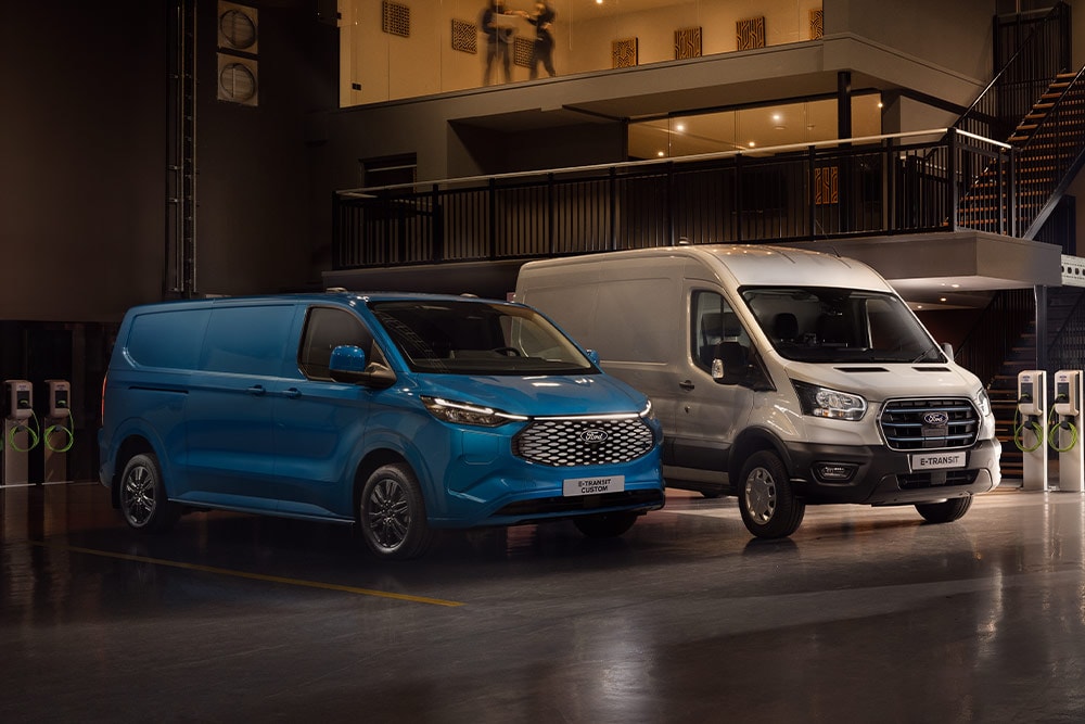 Un Ford E-Transit Custom blu e un Ford E-Transit color argento in ricarica in un parcheggio.