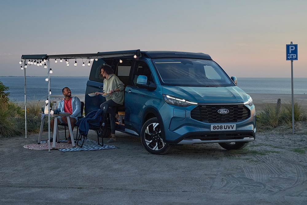 Due campeggiatori si rilassano accanto a un Ford Pro Transit Custom Nugget blu, con un'area di parcheggio sulla spiaggia sullo sfondo.