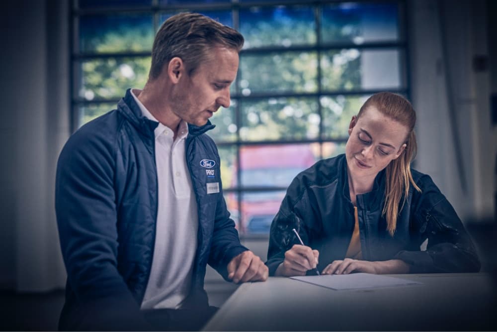 Due dipendenti Ford Pro a un tavolo, uno firma un documento, l'altro osserva in un'officina.
